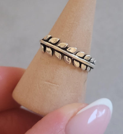 May include: A silver ring featuring a leaf design. The ring is composed of a series of small, oval-shaped leaves arranged in a row along the band. The leaves have a textured appearance, with a dark, oxidized finish. The ring is displayed on a light brown, cone-shaped object.